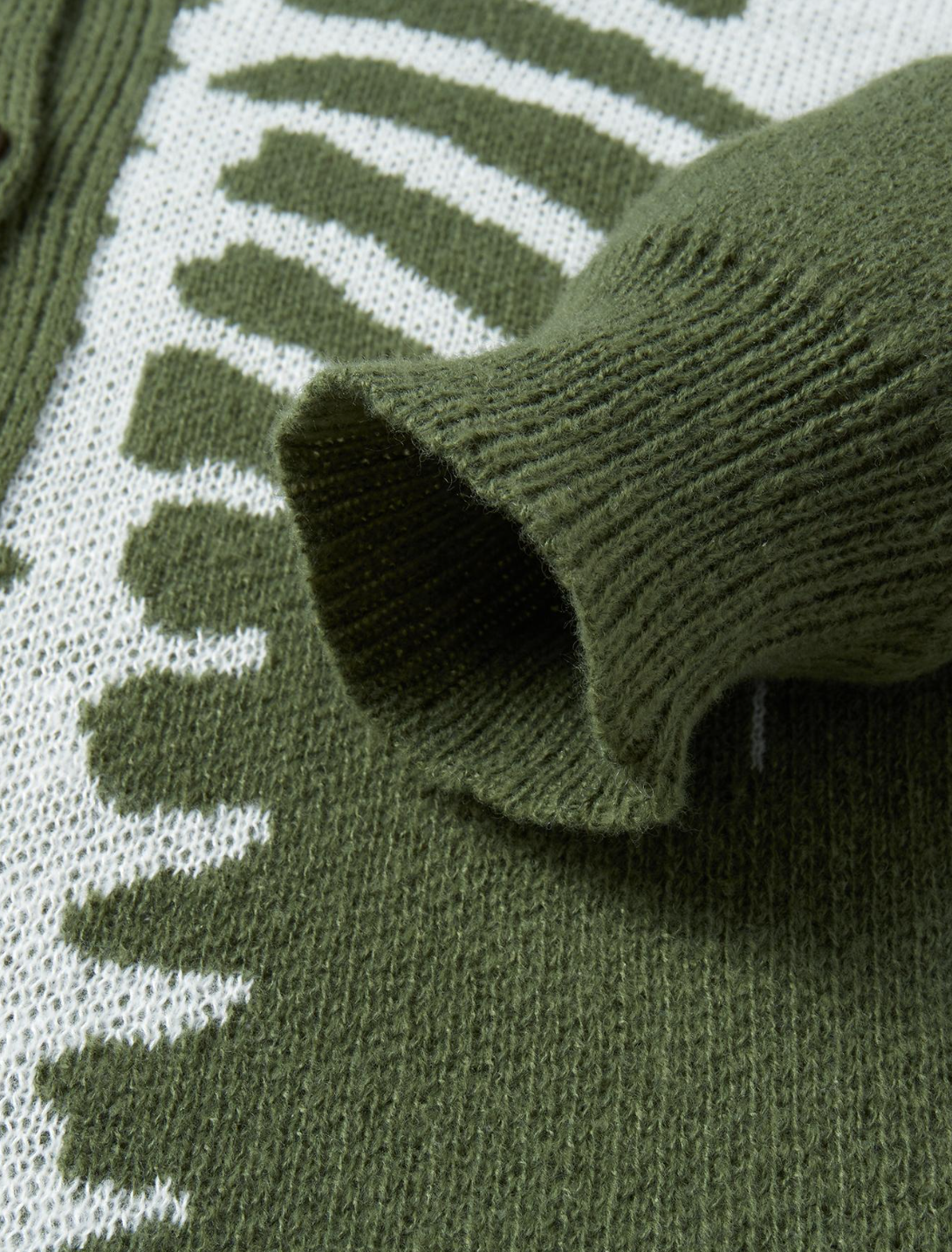 Close-up of an olive green knit sweater with a ribbed cuff and a white knit pattern featuring repeating arch shapes.